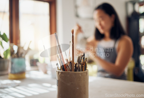 Image of Paintbrush, art and equipment with a jar on a table in a pottery workshop or studio and a designer woman in the background. Zoom, paint and creative with a female potter working on a design
