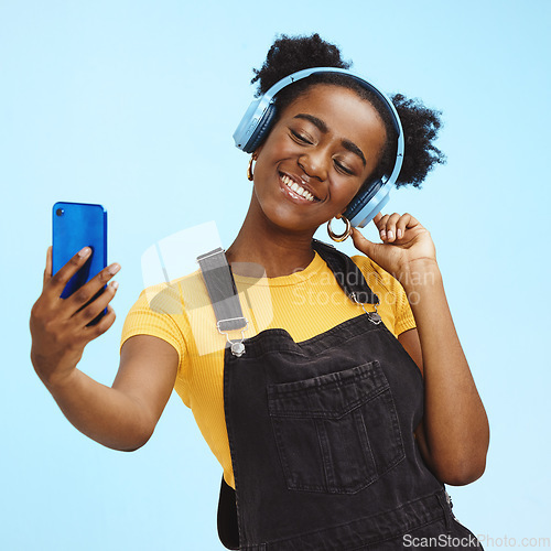 Image of Selfie, smartphone and black woman with headphones, social media and connection on blue studio background. African American female influencer, girl and content creator with smile and online streaming