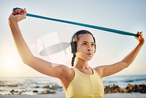 Image of Headphones, beach and woman with band stretching while streaming music, radio or podcast. Sunset, sports and female training with resistance band for health, fitness and wellness outdoors by ocean.