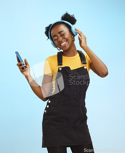 Image of Black woman, phone and music in studio with smile, happiness or listening by blue background. Happy gen z girl, streaming audio or online radio on smartphone for edgy fashion, headphones and backdrop