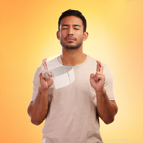 Image of Fingers crossed, wish and man model with orange studio background and hope. Optimism, eyes closed and modern person with a beard hoping for good luck of a young casual male with a tshirt isolated