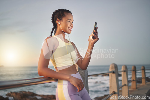 Image of Fitness, beach and woman with smartphone after running at sunset, online and streaming music or podcast in nature for exercise. Runner, relax and scroll social media after run, smile with earphones.