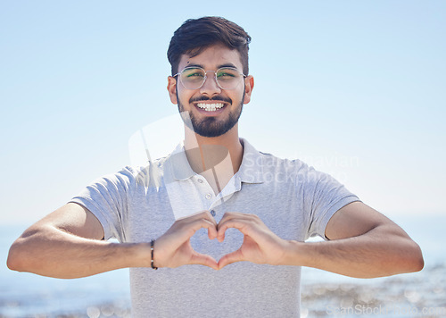 Image of Beach portrait and a man heart hand for summer holiday freedom, happiness and wellness. Happy Indian person enjoying ocean sun with love shape and optimistic smile for travel adventure.