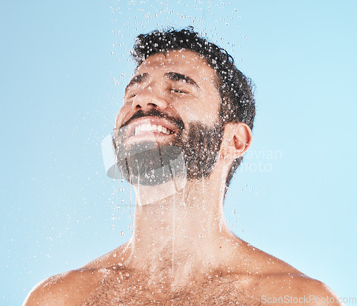 Image of Man, shower and water for cleaning, studio and self care wellness with smile by blue background. Model, skincare and water splash on face, cold and self love for health, body and clean by backdrop