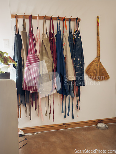 Image of Clothes rack, apron and pottery with a broom in an empty workshop or studio against still life wall for design. Creative, art and clothing with a group of aprons hanging in a small business startup