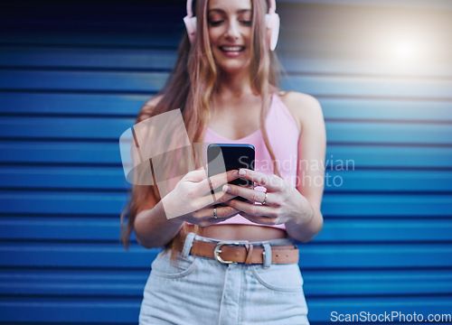 Image of Social media, music and woman reading on a phone with a podcast, communication and app in the city. Headphones, radio and girl typing on a mobile, streaming audio and sound on a blue wall in Germany