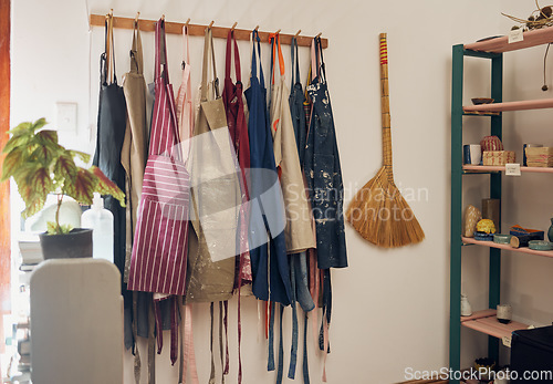 Image of Clothes rack, apron and paint with a broom in an empty workshop or studio against still life wall for design. Creative, art and clothing with a group of aprons hanging in a small business startup
