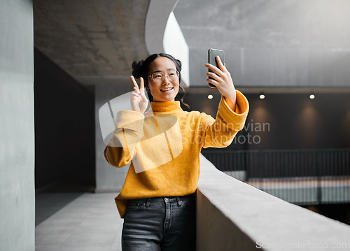 Image of Woman, peace and selfie in office building, smile and happy while on internet, pose and emoji. Asian, girl and business entrepreneur with smartphone for photo, peace sign and hand for profile picture