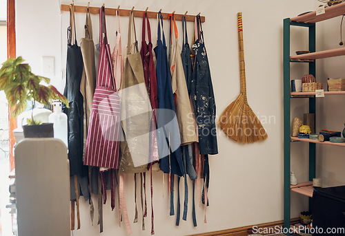 Image of Apron, hanging and art shop with a broom in an empty workshop or studio against still life wall for design. Creative, pottery and clothing with a group of aprons on a rack in a small business startup