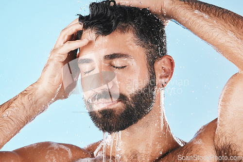 Image of Face, hair care shower and water splash of man in studio isolated on a blue background. Dermatology, water drops and male model washing, cleaning and bathing for healthy skin, skincare and hygiene.