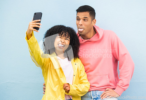 Image of Happy couple, tongue and phone selfie on isolated blue background for social media, city profile picture and travel vlog. Man, black woman and silly faces bonding for on mobile photography technology