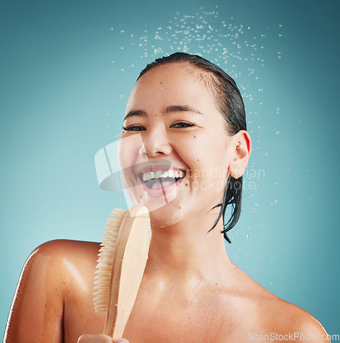 Image of Portrait, bath brush and woman in a shower for cleaning, wellness and skin exfoliate, beauty and grooming in a studio. Skincare, water splash and face of asian model washing, relax on blue background