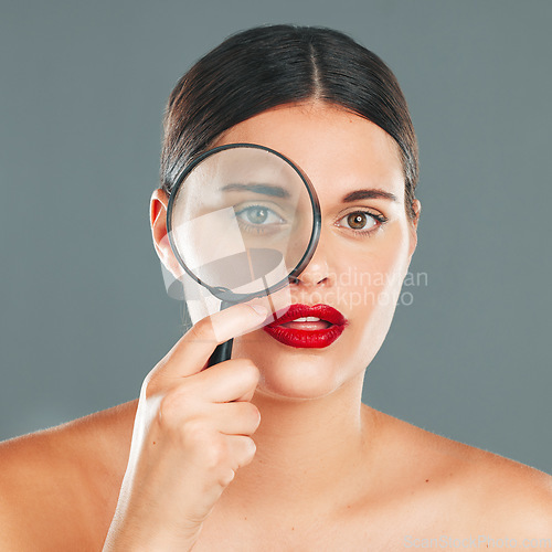 Image of Portrait, magnifying glass and eye with a model woman in studio on a gray background for beauty or skincare. Face, makeup and search with an attractive young woman checking her skin for cleaning