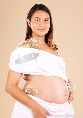Image of Pregnancy, flowers and portrait of a woman in studio with chiffon material on her body holding her stomach. Maternity, prenatal health and pregnant female model with floral posing by beige background