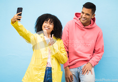 Image of Friends, phone and peace sign for selfie on a blue background for fashion, style or friendship together. Young man and woman smiling and looking at smartphone for photo leaning on a wall in happiness