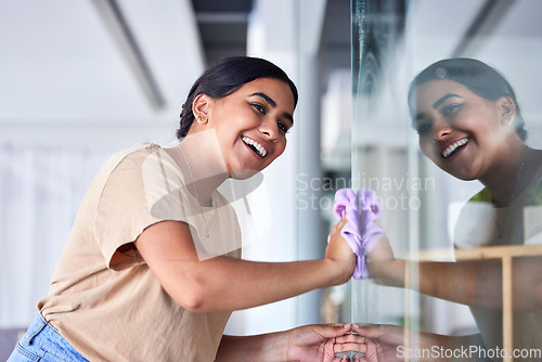 Image of Happy, girl and reflection cleaning window with household cloth and satisfied smile for shiny transparent surface. Happiness, focus and concentration of young Indian woman polishing glass in house.