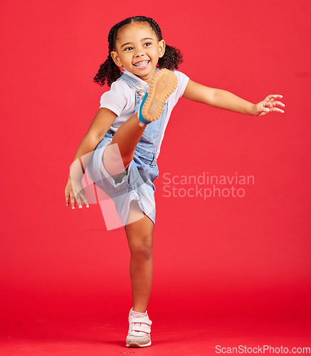 Image of Happy, kick and excited girl in a studio with a casual, stylish and fashion kid outfit. Happiness, smile and young child with cool clothes kicking her leg with excitement isolated by a red background