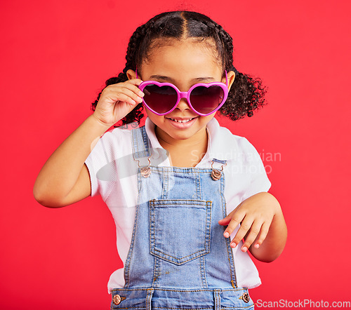 Image of Little girl, fashion and heart sunglasses on isolated red background in children trend, holiday style or cool summer. Smile, happy and kid with glasses for eyes healthcare, wellness or sun protection