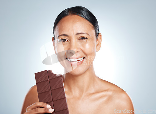 Image of Candy, health and portrait of a woman with chocolate isolated on a blue background in studio. Food, happy and face of an elderly model showing a treat, sugar snack and sweets with smile on a backdrop