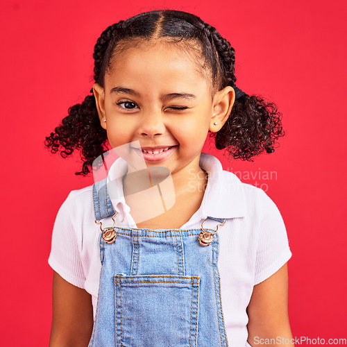 Image of Portrait, wink and a black child on a red background in studio having fun or feeling carefree. Kids, fashion and smile with a happy female child winking inside on a color wall while looking funny