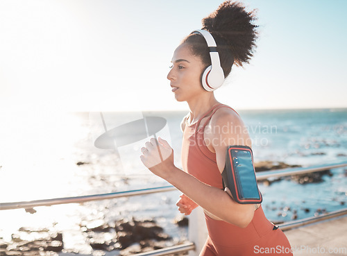 Image of Black woman, fitness and running with headphones and cellphone on arm at the beach in Cape Town. Sporty African American female runner by the ocean coast having a run for cardio training workout