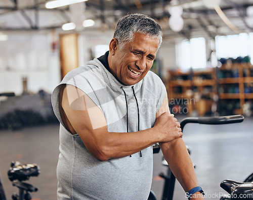 Image of Sports, gym and injury, old man with arm pain, emergency during workout at fitness studio. Health, wellness and inflammation, senior person with hand on muscle cramps while training on exercise bike.