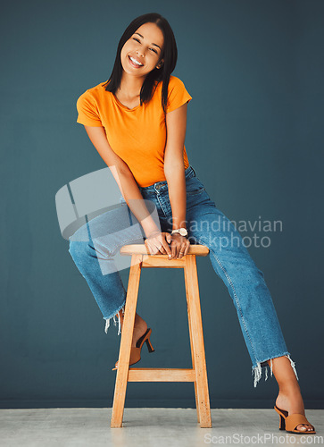 Image of Black woman, smile and portrait of a young model sitting on a stool in a studio. Casual fashion, happiness and youth of a female feeling relax, calm and happy on a chair isolated by a blue wall