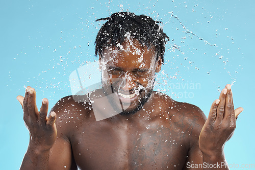 Image of Water splash, cleaning and hygiene with a model black man in studio on a blue background for hydration. Bathroom, skincare and wellness with a young male wahsing his face for natural skin treatment