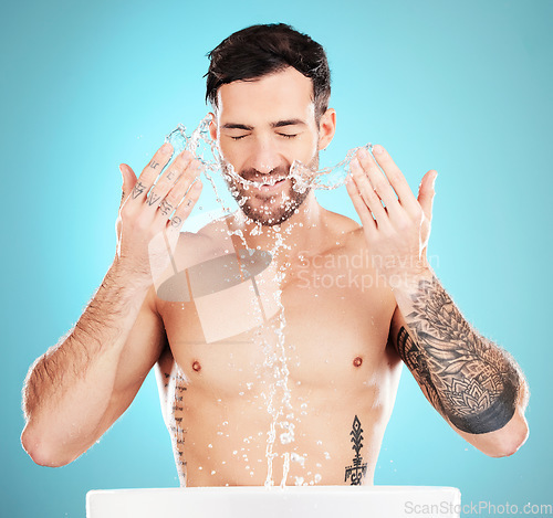 Image of Water splash, skincare and man cleaning face, morning detox treatment isolated on blue background. Facial hygiene, male model with tattoo and grooming for health, wellness and skin care in studio.