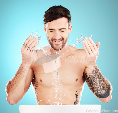 Image of Water splash, sink and skincare of a man cleaning face for wellness, self care and dermatology. Liquid, model ad blue background in a studio with a young man with tattoo doing treatment for beauty