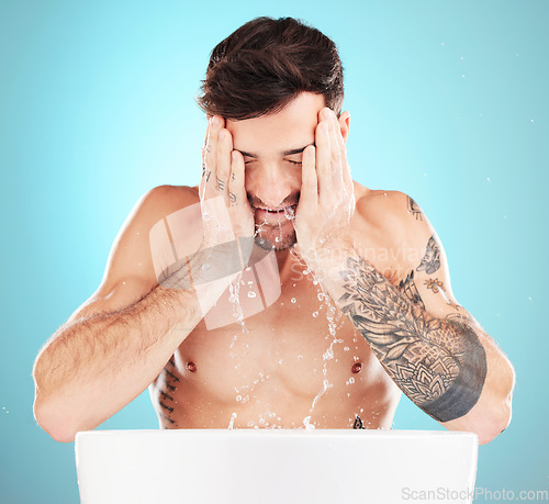 Image of Water, skincare and man with cleaning face, morning skin treatment isolated on blue background. Facial hygiene, splash and male model with tattoo grooming for health, wellness and beauty in studio.