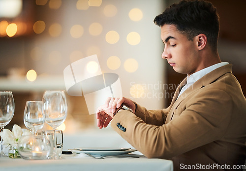 Image of Time check, watch and late date of a man at a restaurant table on valentines day alone. Bokeh lights, night and suit of a person waiting for dinner and looking at his smartwatch on anniversary