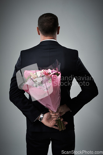 Image of Valentines day, surprise and flowers of a man in a suit with love present behind back. Flower gift, suit and male model in a isolated gray background studio feeling romantic for a formal date