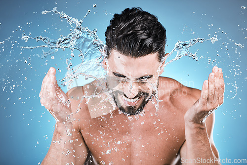 Image of Water splash, hands and a man model washing his face in studio on a blue background for hygiene or hydration. Bathroom, skincare and cleaning with a handsome young male splashing his skin for beauty