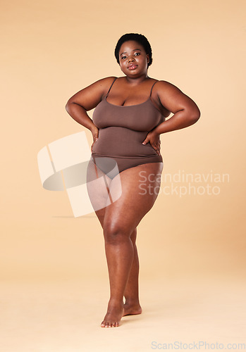 Image of Portrait, wellness and plus size with a model black woman posing hands on hips in studio on a beige background. Beauty, underwear and natural with a female sitting on a stool for body positivity