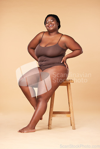 Image of Portrait, wellness and plus size with a model black woman posing on a chair in studio on a beige background. Beauty, underwear and natural with a female sitting on a stool for body positivity