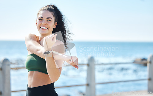 Image of Portrait, fitness and stretching arm by woman at beach for running, exercise or cardio on blue sky background. Face, stretch or workout run by girl at ocean training, happy or relax warm up routine