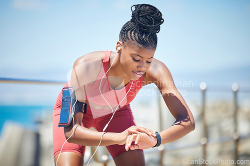Image of Fitness, smartwatch and runner time of a black woman by the sea doing exercise and running workout. Outdoor, run tracker app and mobile of a athlete with headphones by the ocean listening to audio