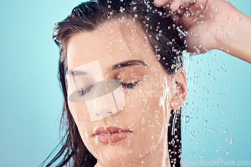 Image of Water splash, hair care and face of woman in shower in studio isolated on a blue background. Beauty, eyes closed and young female model washing, cleaning or bathing for hygiene, skincare and health.