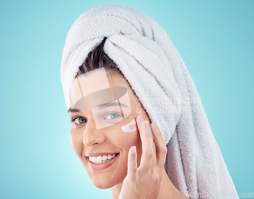 Image of Face, towel and woman with skincare cream in studio isolated on a blue background. Portrait, dermatology and happy female model apply lotion cosmetics, creme or facial moisturizer for healthy skin.