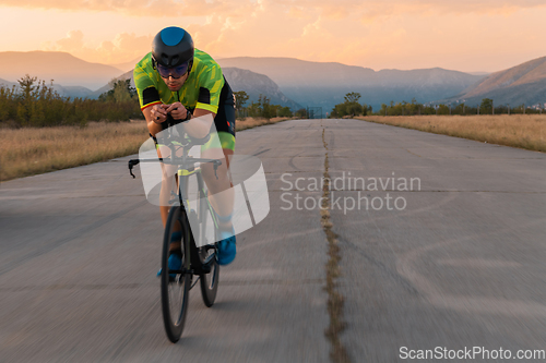 Image of Triathlete riding his bicycle during sunset, preparing for a marathon. The warm colors of the sky provide a beautiful backdrop for his determined and focused effort.