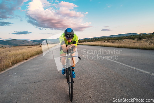 Image of Triathlete riding his bicycle during sunset, preparing for a marathon. The warm colors of the sky provide a beautiful backdrop for his determined and focused effort.