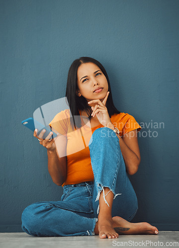 Image of Phone, thinking and social media with a woman in studio on a blue background, sitting on the floor. Mobile, contact or idea and an attractive young female posing with her finger on her chin