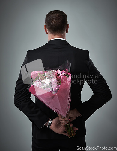 Image of Valentines day, surprise bouquet and flowers of a man in a suit with love present behind back. Flower gift, suit and male model in a isolated gray background studio feeling romantic for a formal date