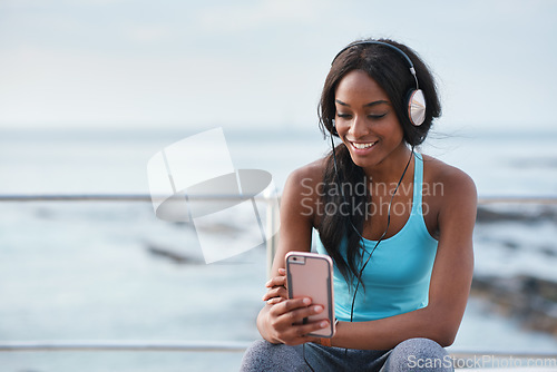 Image of Black woman, phone and headphones at beach, sitting and smile for video, music or funny podcast to relax. Runner, rest and smartphone with meme, social media app with happiness on outdoor adventure