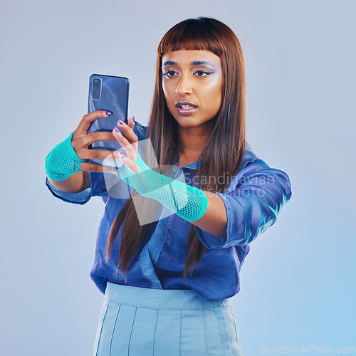 Image of Fashion, selfie and woman with smartphone and cyberpunk neon clothing isolated on blue background. Social media, future and trendy gen z influencer from India with phone in studio for profile picture