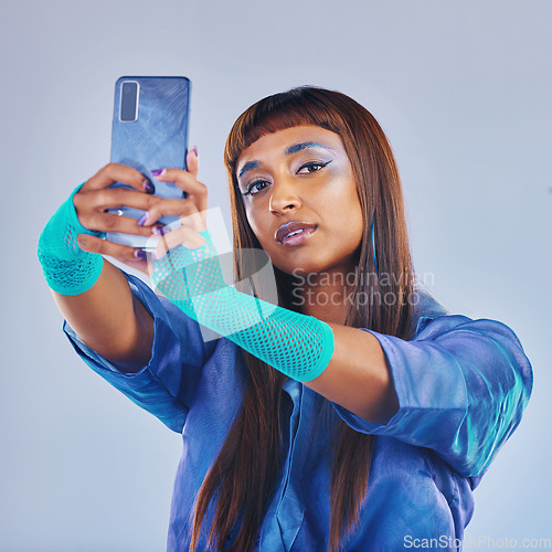 Image of Fashion, selfie and woman with smartphone and cyberpunk neon clothing isolated on blue background. Social media, future and trendy gen z influencer from India with phone in studio for profile picture