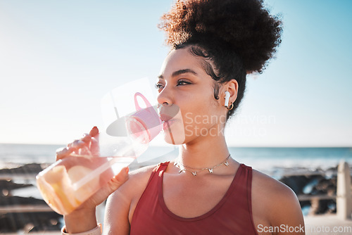 Image of Fitness black woman drinking water on beach for exercise, listening to music and cardio training in blue sky lens flare. Liquid bottle for diet, goals and tired sports runner or USA person by ocean