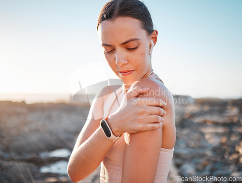 Image of Beach, fitness and woman holding shoulder pain, standing on rocks at ocean. Nature, sports and arm injury during workout for health and wellness, muscle trauma during stretching exercise in morning.