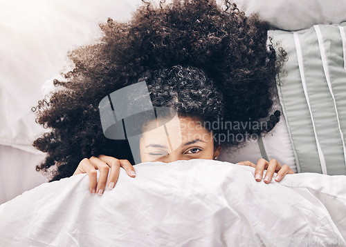 Image of Bed, wink portrait and black woman in the morning after sleep and rest at home with blanket. Eyes, house and wake up happiness of a young person hiding face under the bedroom covers on a pillow